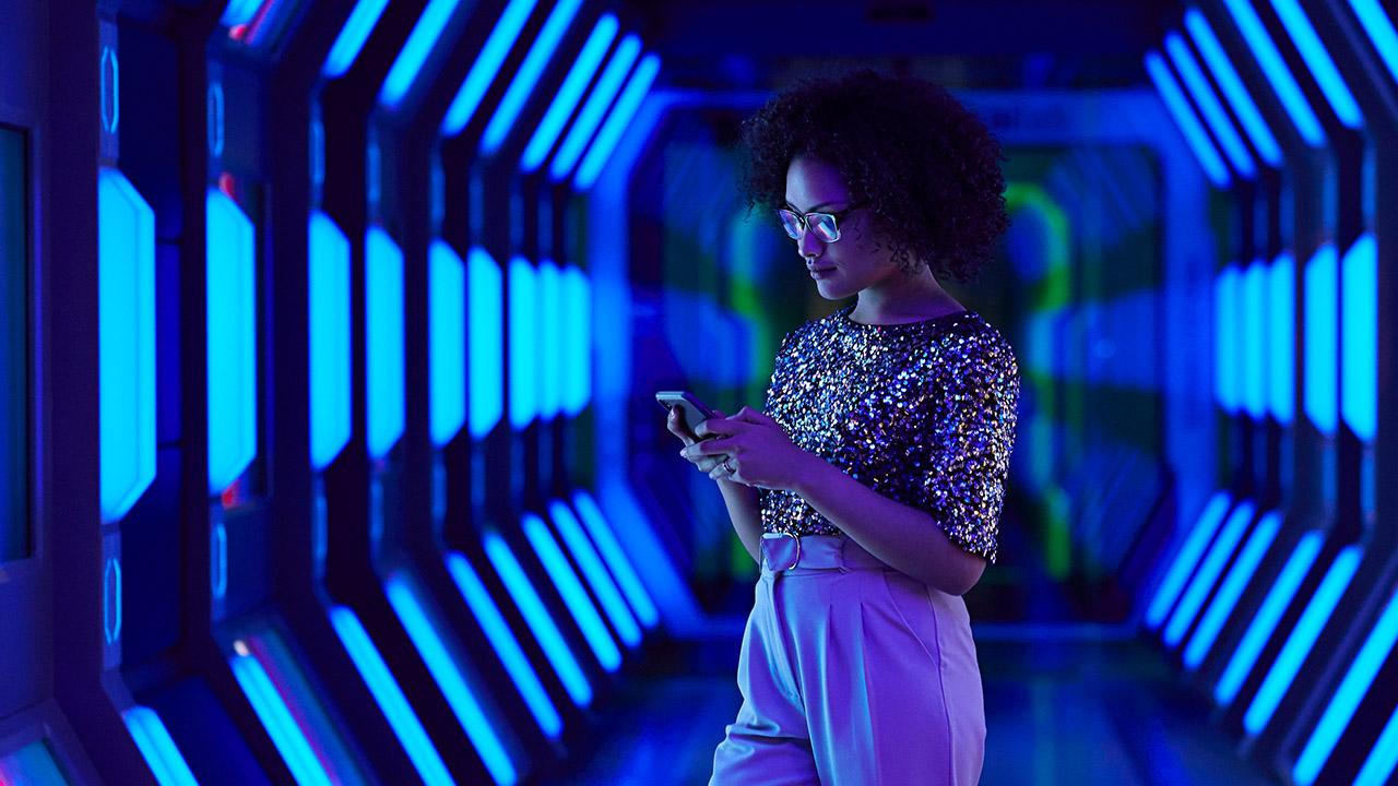 Frau in glitzerndem Oberteil steht in einem futuristisch beleuchteten Korridor und schaut auf ihr Smartphone.
