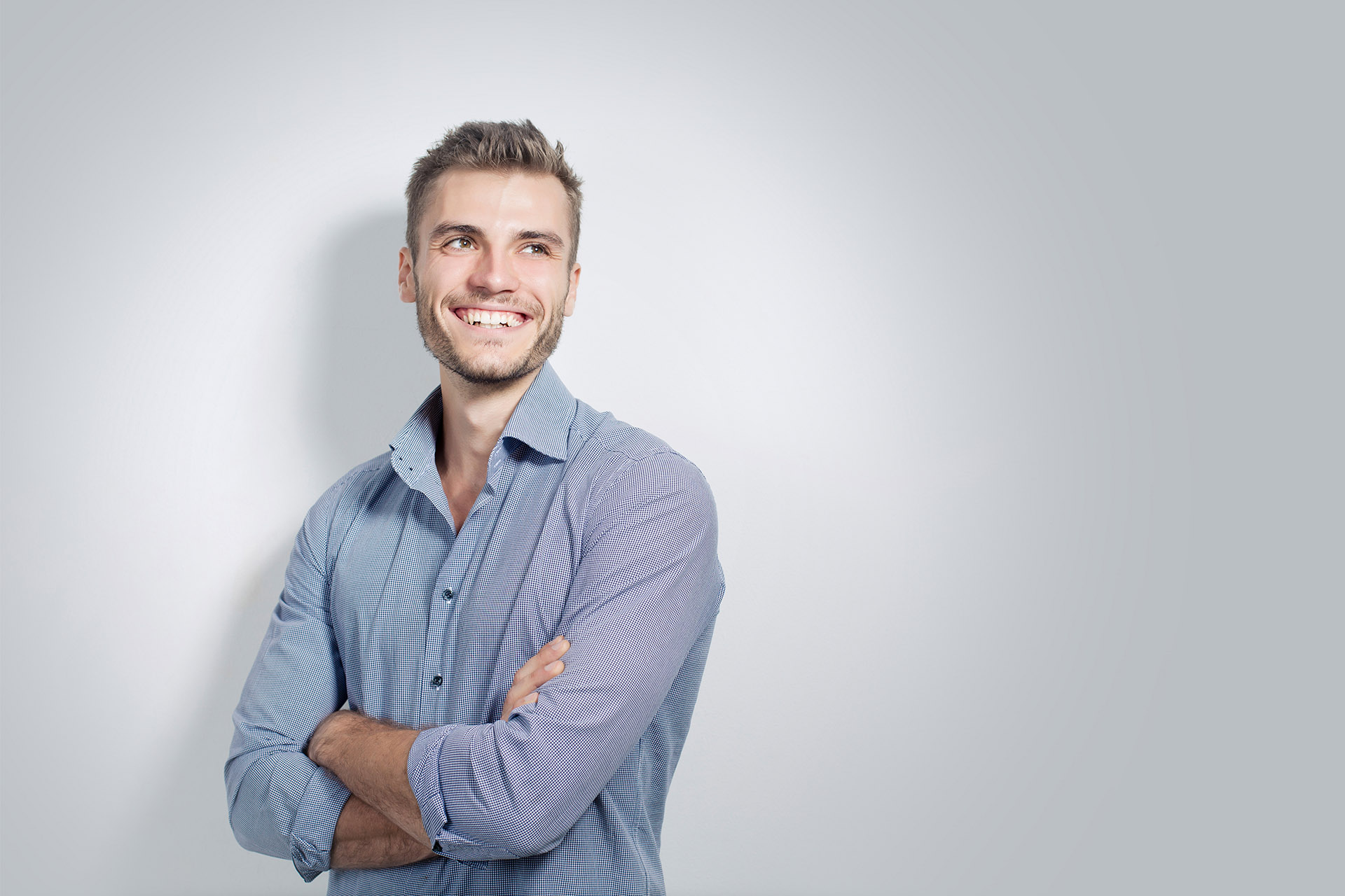 Lächelnder Mann in einem blauen Hemd steht mit verschränkten Armen vor einem hellen Hintergrund, blickt zur Seite.